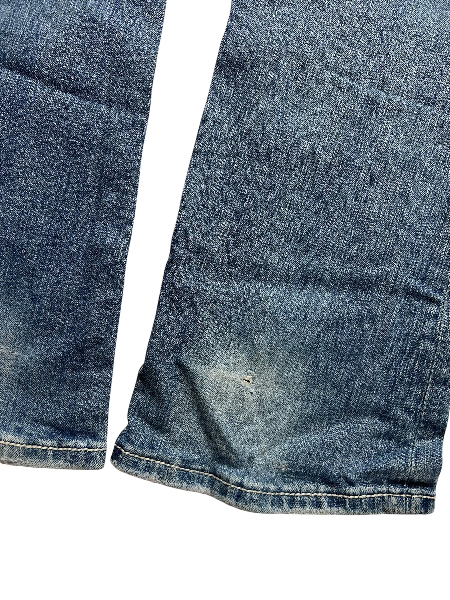 Die Nahaufnahme der Miss Me Low Rise Boot Jeans (Größe 26, S) von ZAZA resell zeigt verblichenen blauen Denim und einen kleinen Riss in der Nähe des Saums. Die Jeans liegt flach auf einem weißen Hintergrund. Labelgröße und Bundweite sind auf Anfrage erhältlich.