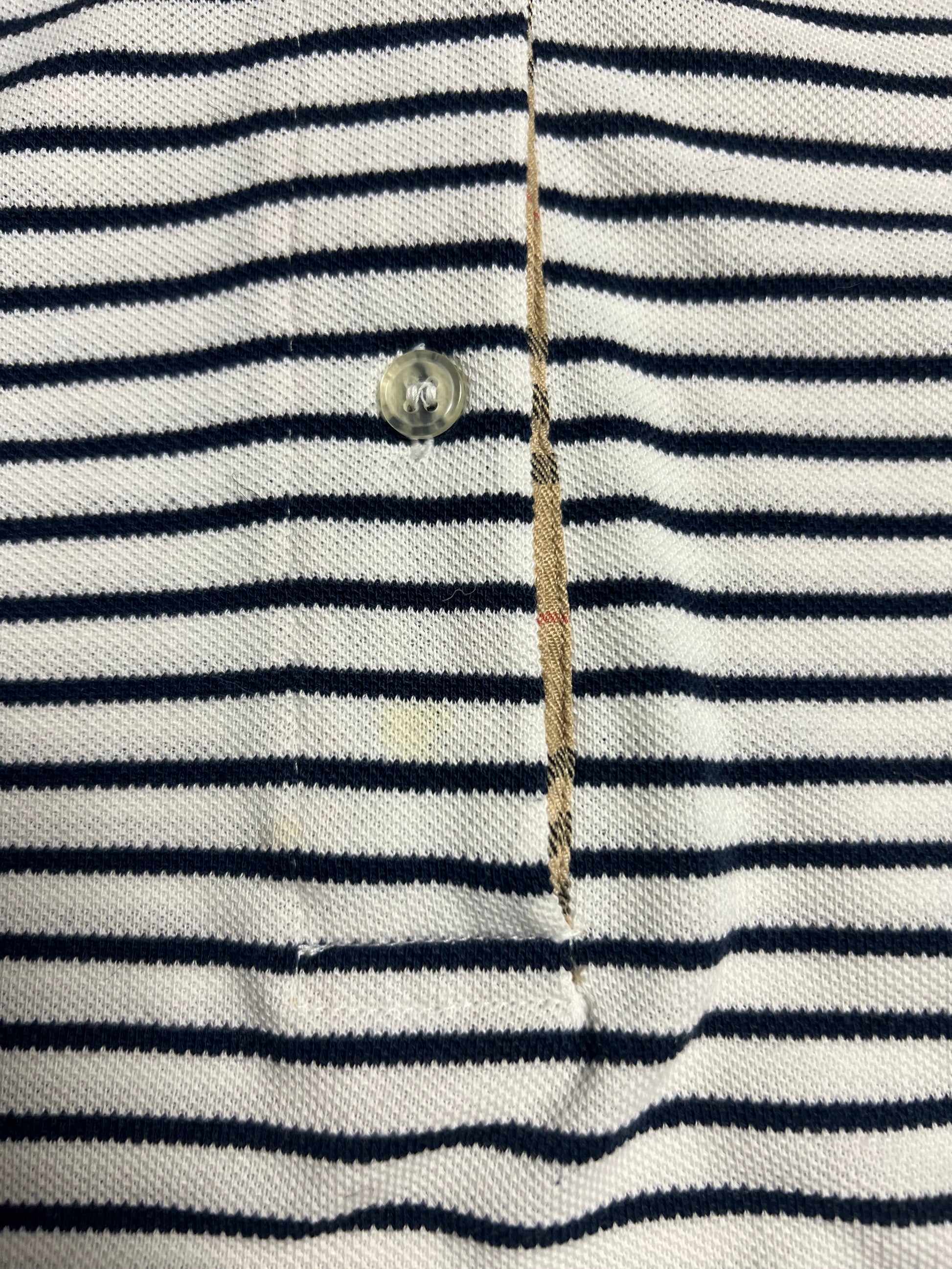 Nahaufnahme eines Burberry Polo Shirts | S von ZAZA resell, weiß mit schwarzen Querstreifen. Mit durchsichtigem Plastikknopf, aufgeknöpfter Knopfleiste, sichtbaren Nähten und beigem Innenfutter. Zustand 8/10. Maße in Zentimetern auf Anfrage.