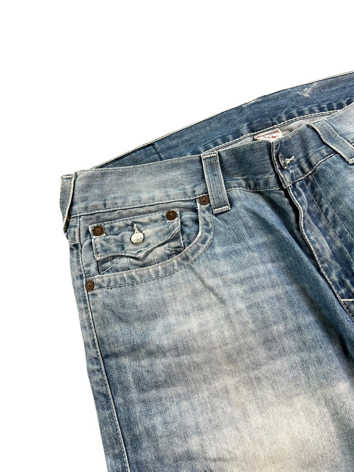 Eine Nahaufnahme der True Religion Straight Jeans von ZAZA resell in Größe 36 (XL). Sie zeigt abgenutzten, verblichenen blauen Denim, eine Vordertasche mit Metallknopf und Nieten sowie das Größenetikett auf dem Bund vor einem weißen Hintergrund.
