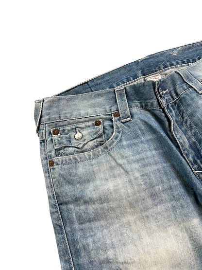 Eine Nahaufnahme der True Religion Straight Jeans von ZAZA resell in Größe 36 (XL). Sie zeigt abgenutzten, verblichenen blauen Denim, eine Vordertasche mit Metallknopf und Nieten sowie das Größenetikett auf dem Bund vor einem weißen Hintergrund.