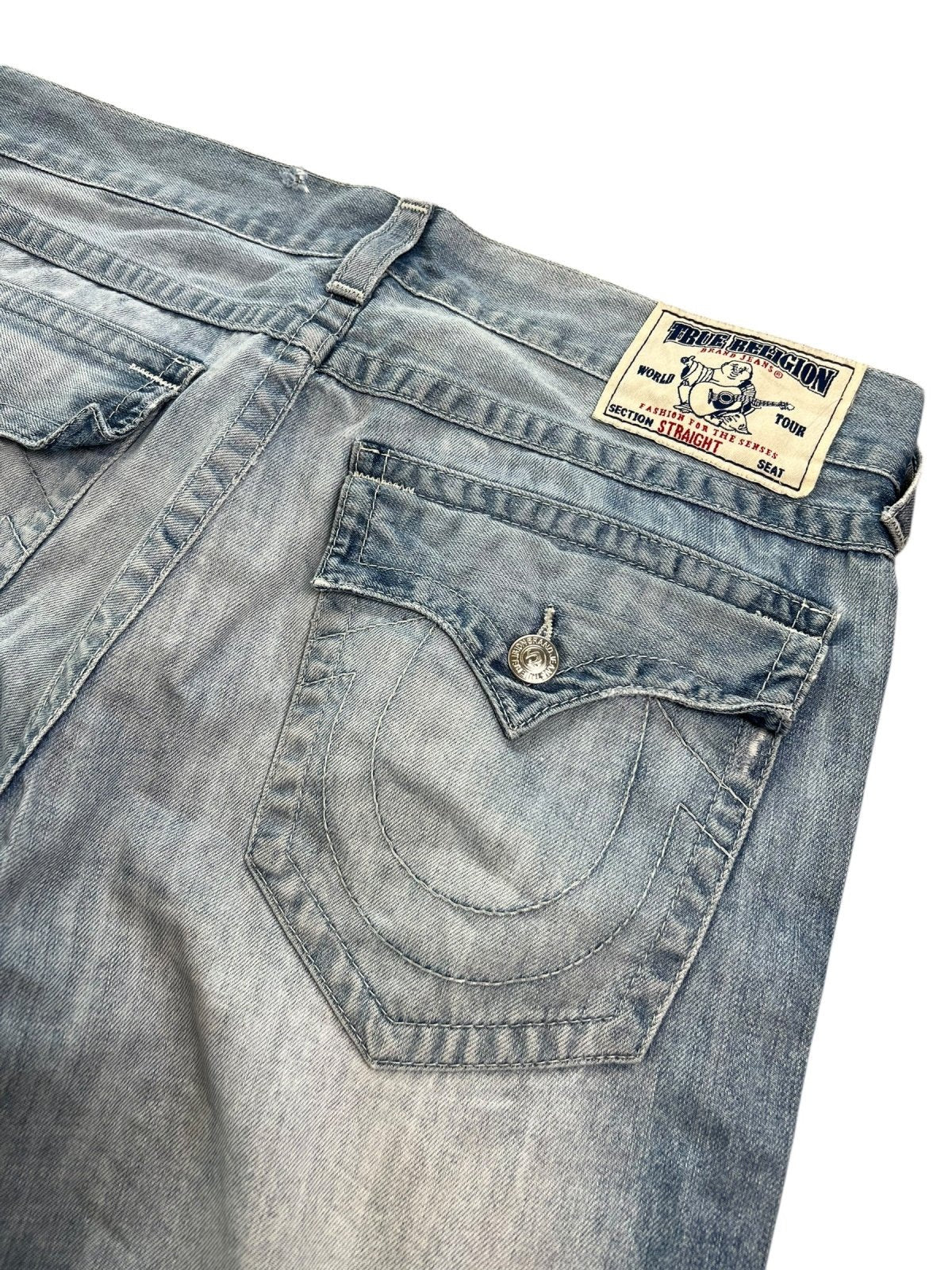 Eine Nahaufnahme der True Religion Straight | 36 (XL) Jeans von ZAZA resell zeigt die Gesäßtasche mit detaillierten Nähten, die geknöpfte Klappe und das charakteristische Gitarrenlabel am Bund, das Merkmale wie Bundweite und Außenbeinlänge hervorhebt.