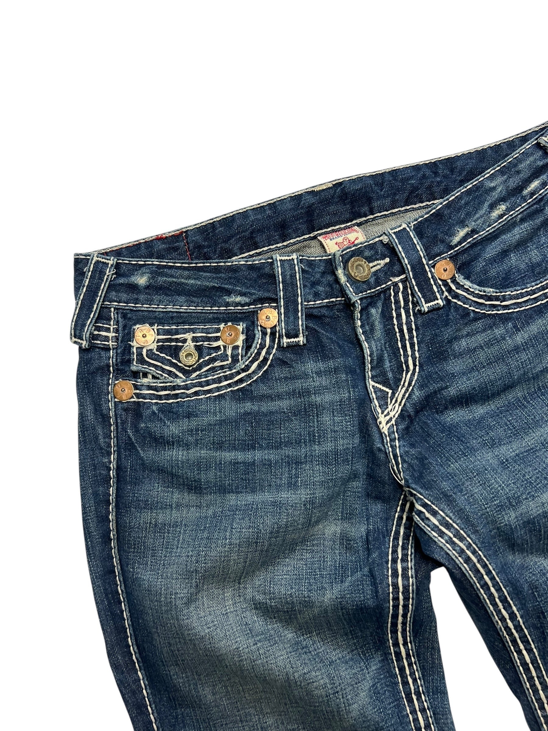 Eine Nahaufnahme der True Religion Joey Big T - Low Rise Boot Jeans (28/M) aus dem ZAZA-Wiederverkauf. Sie zeigt blauen Denim mit auffälligen weißen Nähten, Metallnieten an mehreren Taschen, einen leicht ausgeblichenen Look, Bundweite 40 markiert und den oberen vorderen Bereich teilweise gefaltet.