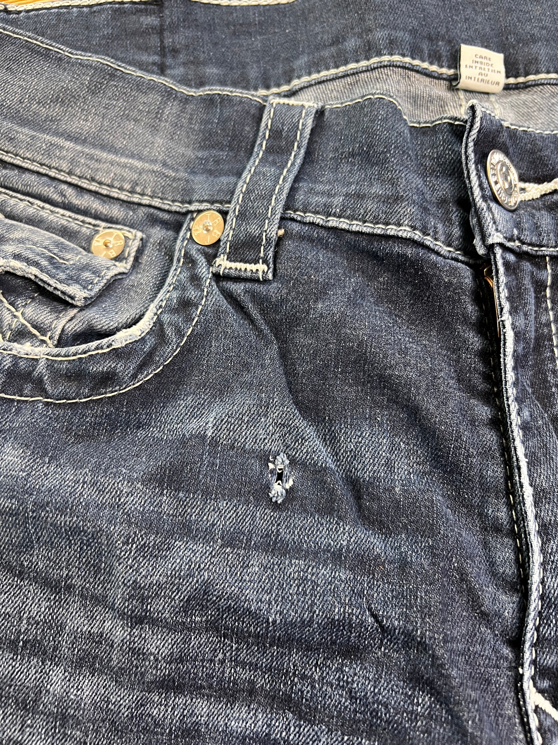 Eine Nahaufnahme der True Religion Straight Jeans (W36 x L36) von ZAZA Resell zeigt die Vordertasche, den Knopf, die Gürtelschlaufe und eine kleine Reparatur in der Nähe der Tasche; der Stoff ist abgenutzt und leicht verblasst – „Zustand“ weist auf sanften Gebrauch hin.