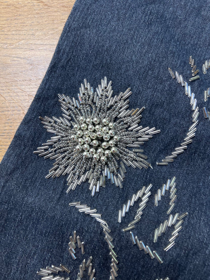 Nahaufnahme des Denim-Schuhs „Edith Mid Rise Boot“ (B26 x L32) von ZAZA. Er zeichnet sich durch eine auffällige, silberne Blumenverzierung mit runden Perlen und röhrenförmigen Blütenblättern in der Nähe der Beinöffnung aus. Im Hintergrund ist eine Holzoberfläche zu sehen.