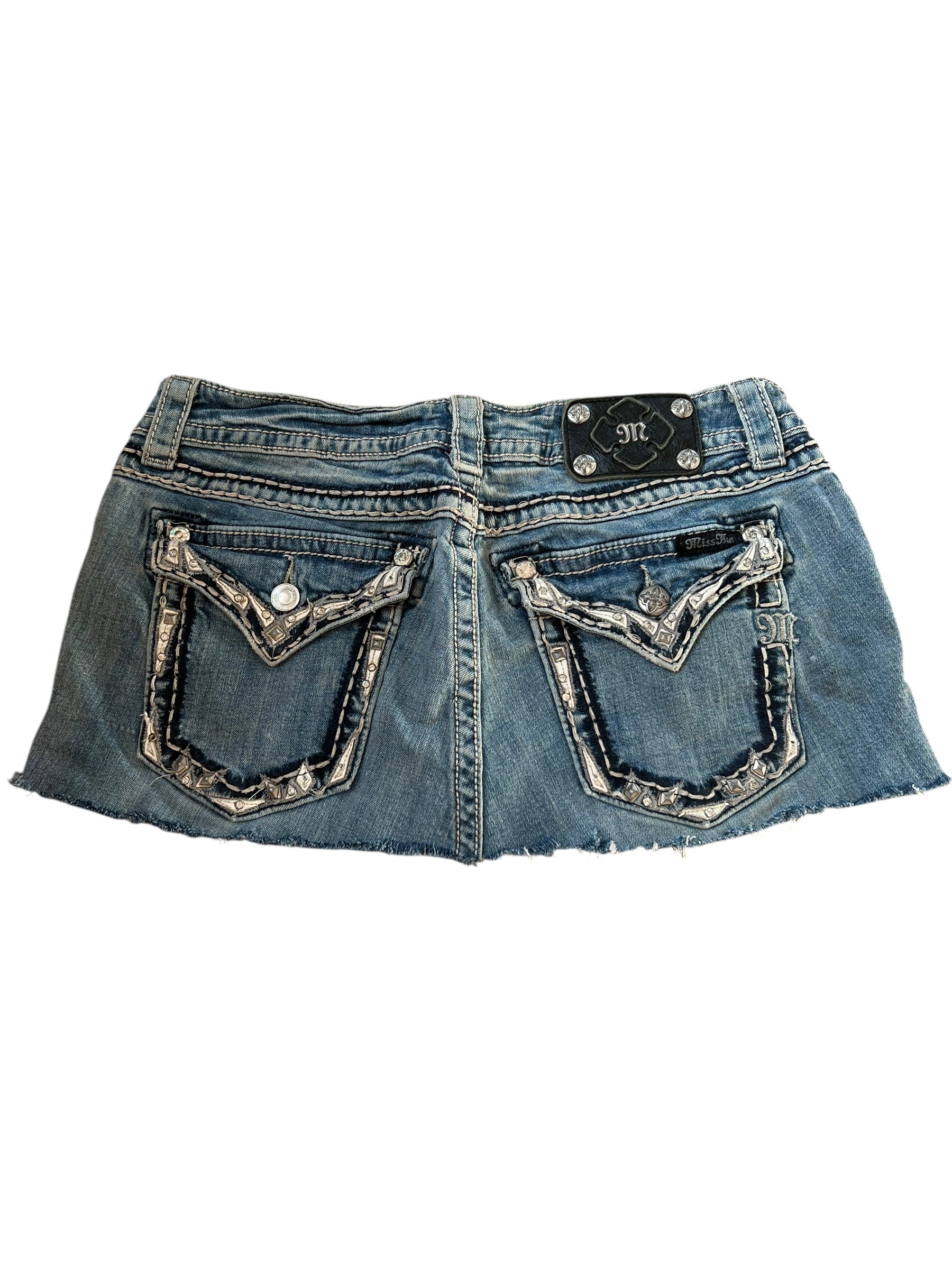 Der Miss Me Skirt | 31 (M) von ZAZA resell ist ein Denim-Minirock mit ausgefranstem Saum, aufgesetzten Gesäßtaschen, Metallknöpfen und einem schwarzen Bundaufnäher mit silbernen Akzenten. Labelgröße 31, Bundweite 38 für eine individuelle Rock-Passform.