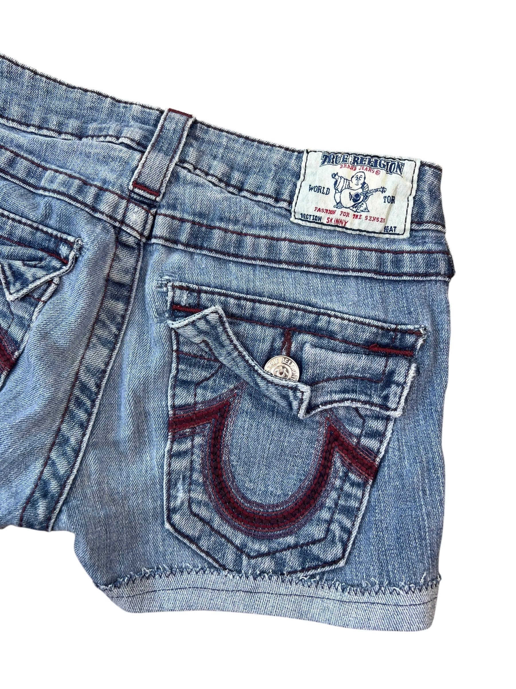 Nahaufnahme der blauen True Religion Shorts aus Denim (Größe 25/XS) von ZAZA resell mit Gesäßtasche mit Klappe, roten Nähten, Metallknopf und einem weißen Etikett mit einem Gitarristen über der Tasche.