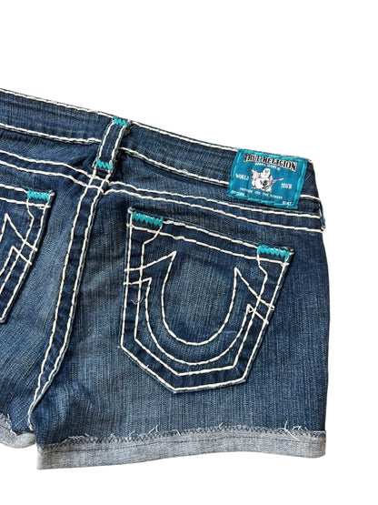 Eine Nahaufnahme der blauen True Religion Shorts (Größe 30/M) von ZAZA resell mit weißen Nähten, ausgefranstem Saum, hufeisenförmigem Detail an der Gesäßtasche und einem blauen Markenetikett über der rechten Tasche. Außenbeinlänge 26 cm.