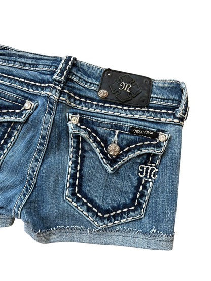 Nahaufnahme der Miss Me Shorts | 27 (S) von ZAZA resell aus blauem Denim mit Bundweite 35, weißen Kontrastnähten, silbernen Knöpfen, einer bestickten Gesäßtasche und einem schwarzen Lederpatch mit M-Logo in der Nähe des Bundes.