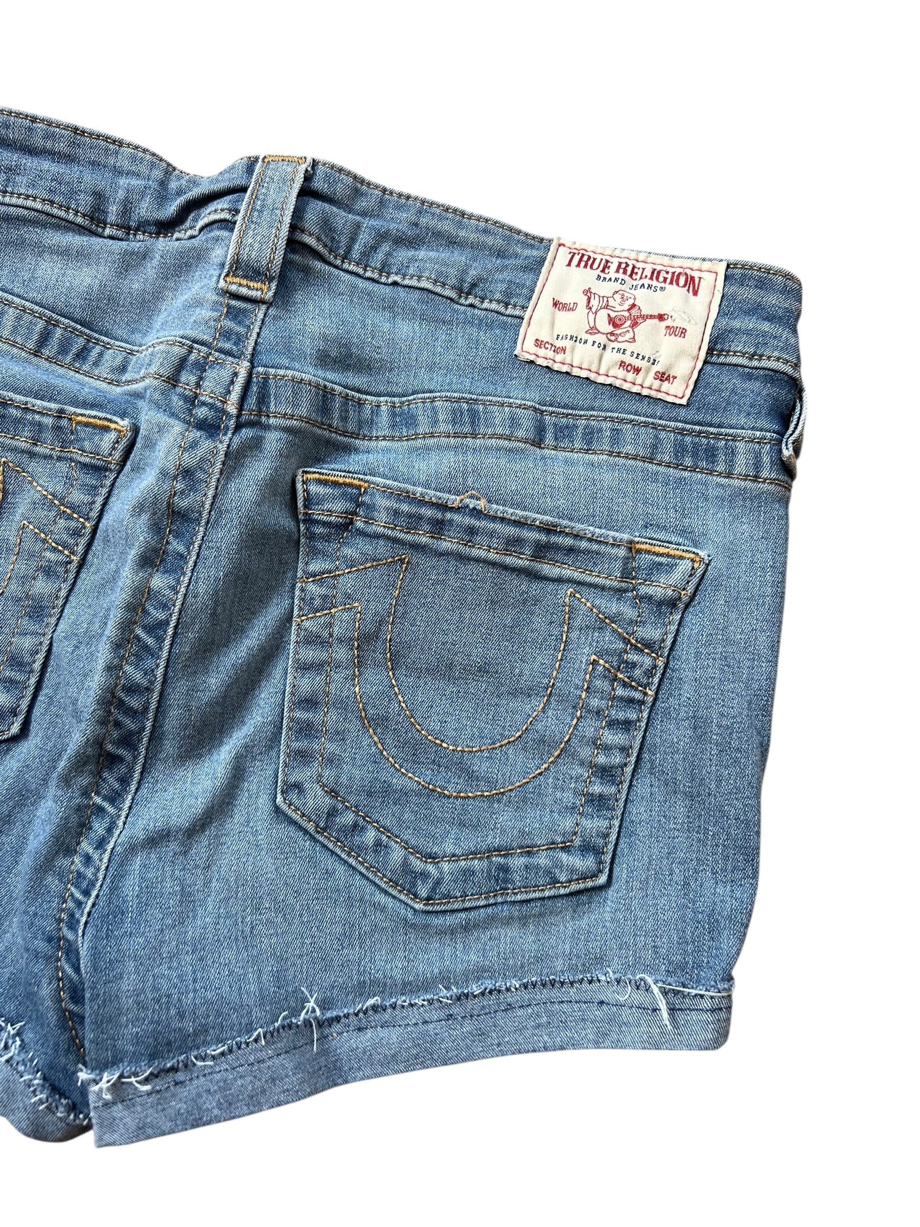 Eine Nahaufnahme der blauen True Religion High Waist Shorts (Größe 30, M) von ZAZA resell mit ausgefranstem Saum, aufgenähtem Hufeisen auf der Gesäßtasche und weißem Markenlabel am Bund.