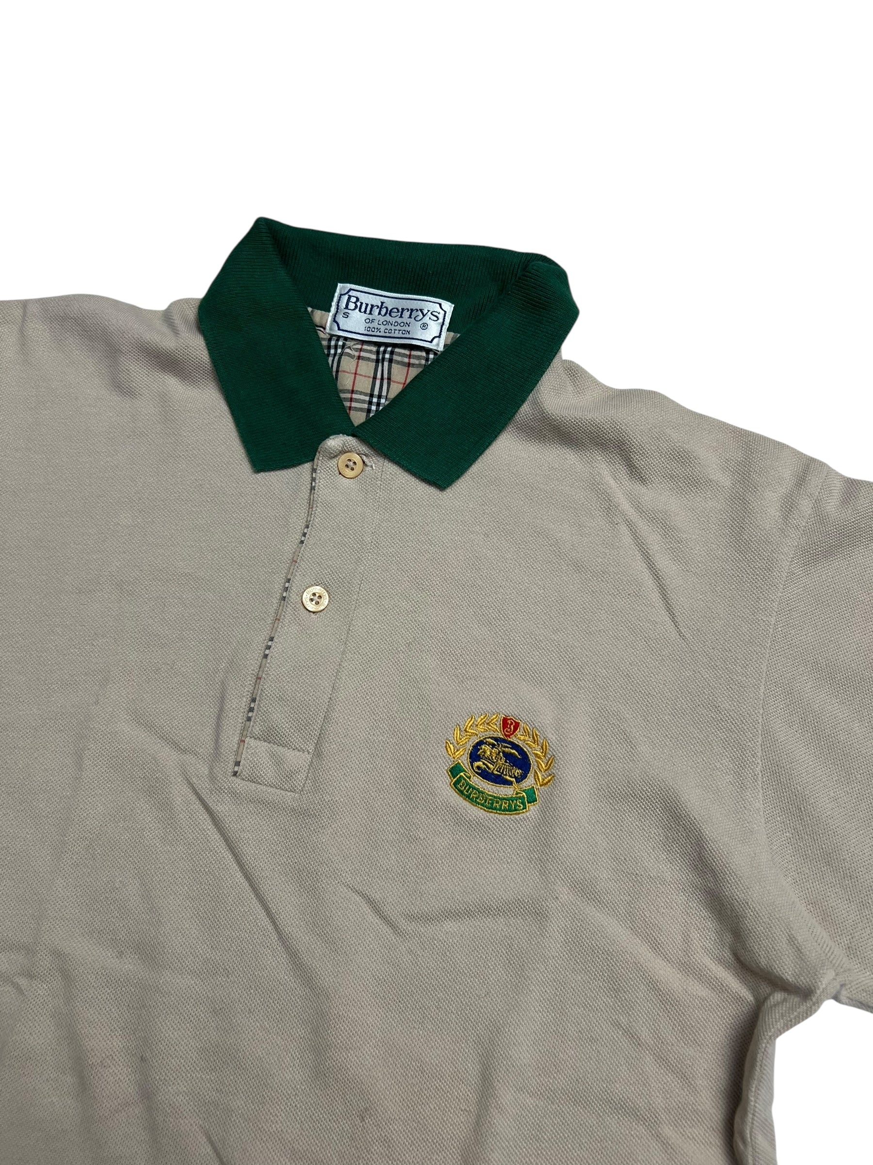 Burberry Polo Shirt | S von ZAZA resell in beige mit grünem Kragen, zwei Knöpfen und gesticktem Wappenlogo auf der linken Brust. Innenkragen mit Burberry-Etikett und Signatur-Karo. Zustand 9/10. Maße in cm auf Anfrage.