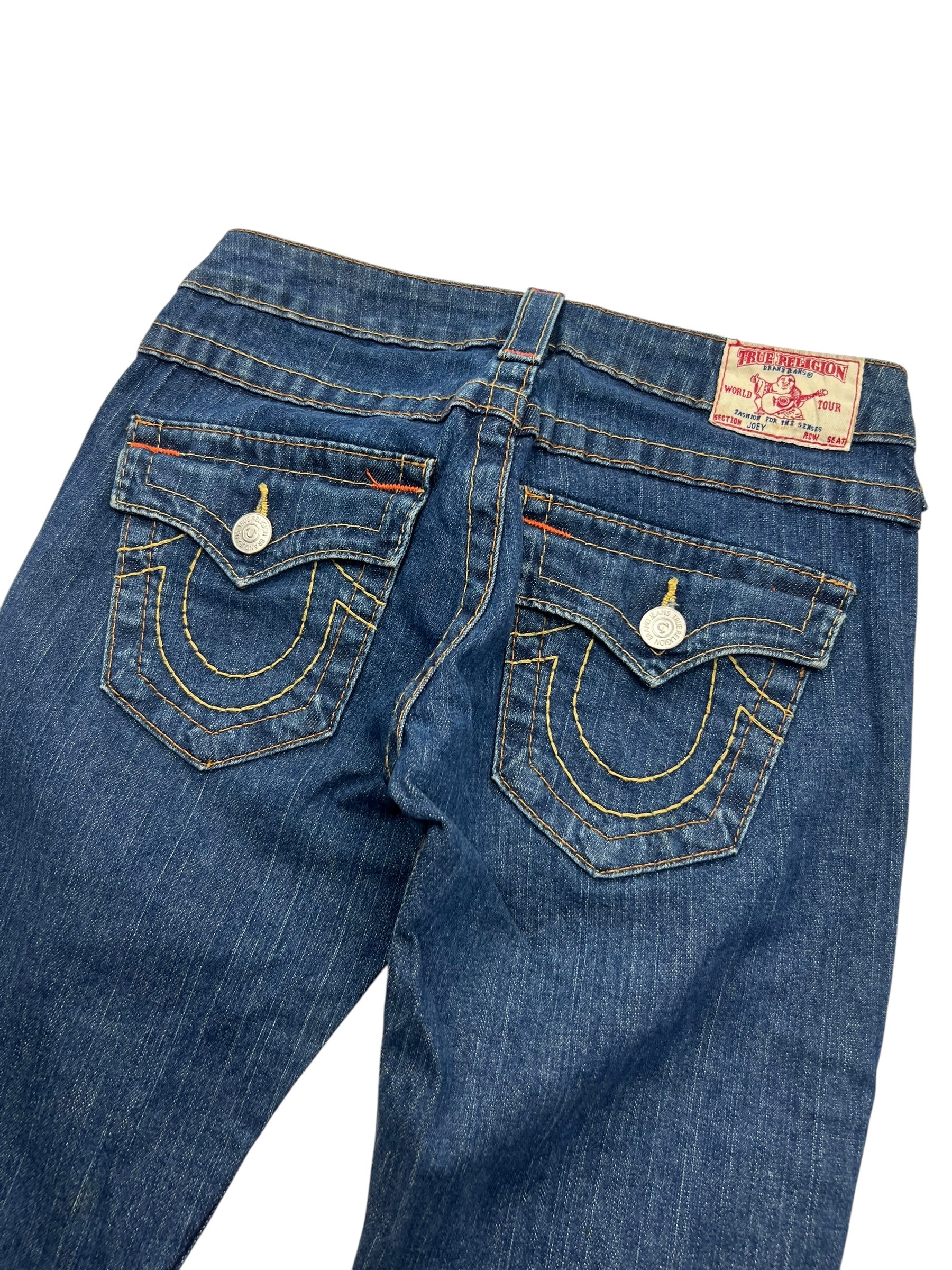 Eine Nahaufnahme der Rückseite der True Religion Joey - Low Rise Boot Jeans von ZAZA resell, mit Pattentaschen mit Hufeisennaht, silbernen Knöpfen, einem rot-weißen Label am Bund; Bundweite 36, Außenbeinlänge 97 cm.