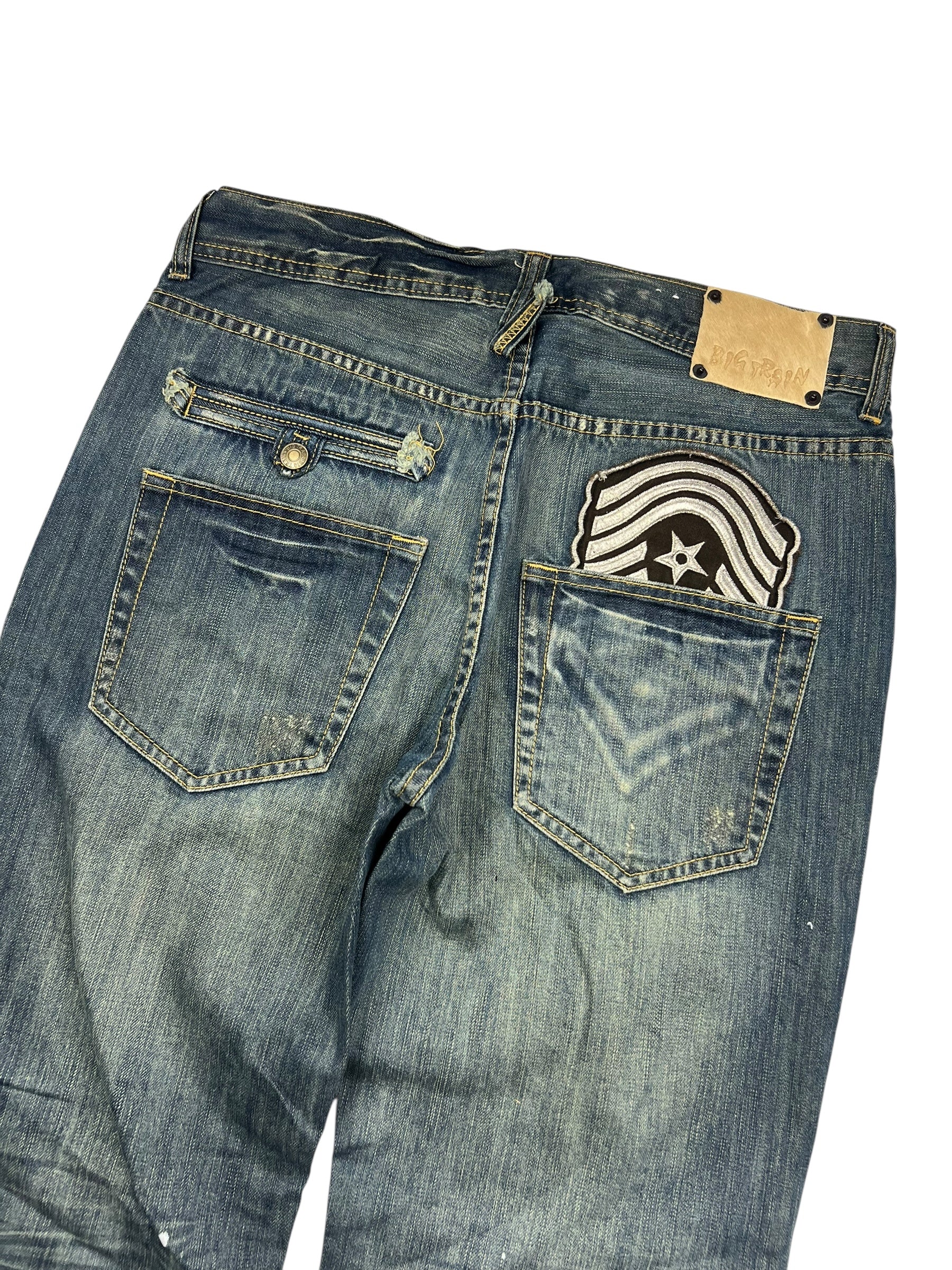 Ein Paar Big Train Boot Jeans in M/L von ZAZA resell, abgebildet von hinten mit verblasstem blauem Denim, einem Stern- und Streifenaufnäher über einer Tasche, einem Lederetikett am Bund und einer Bundweite plus Außenbeinlänge für eine ideale Passform.