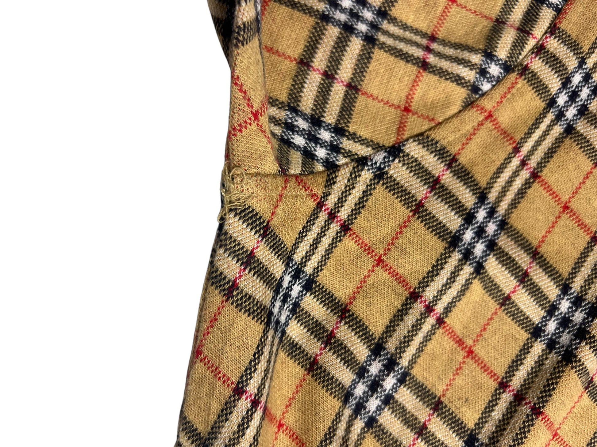 Nahaufnahme eines hellbraunen Burberry-Poloshirts (Größe S) mit einem karierten Muster aus schwarzen, weißen und roten Linien. Das Bild zeigt eine Naht und eine Falte, die auf Kleidungsdetails hinweisen. Marke: ZAZA resell. Zustand 9/10.