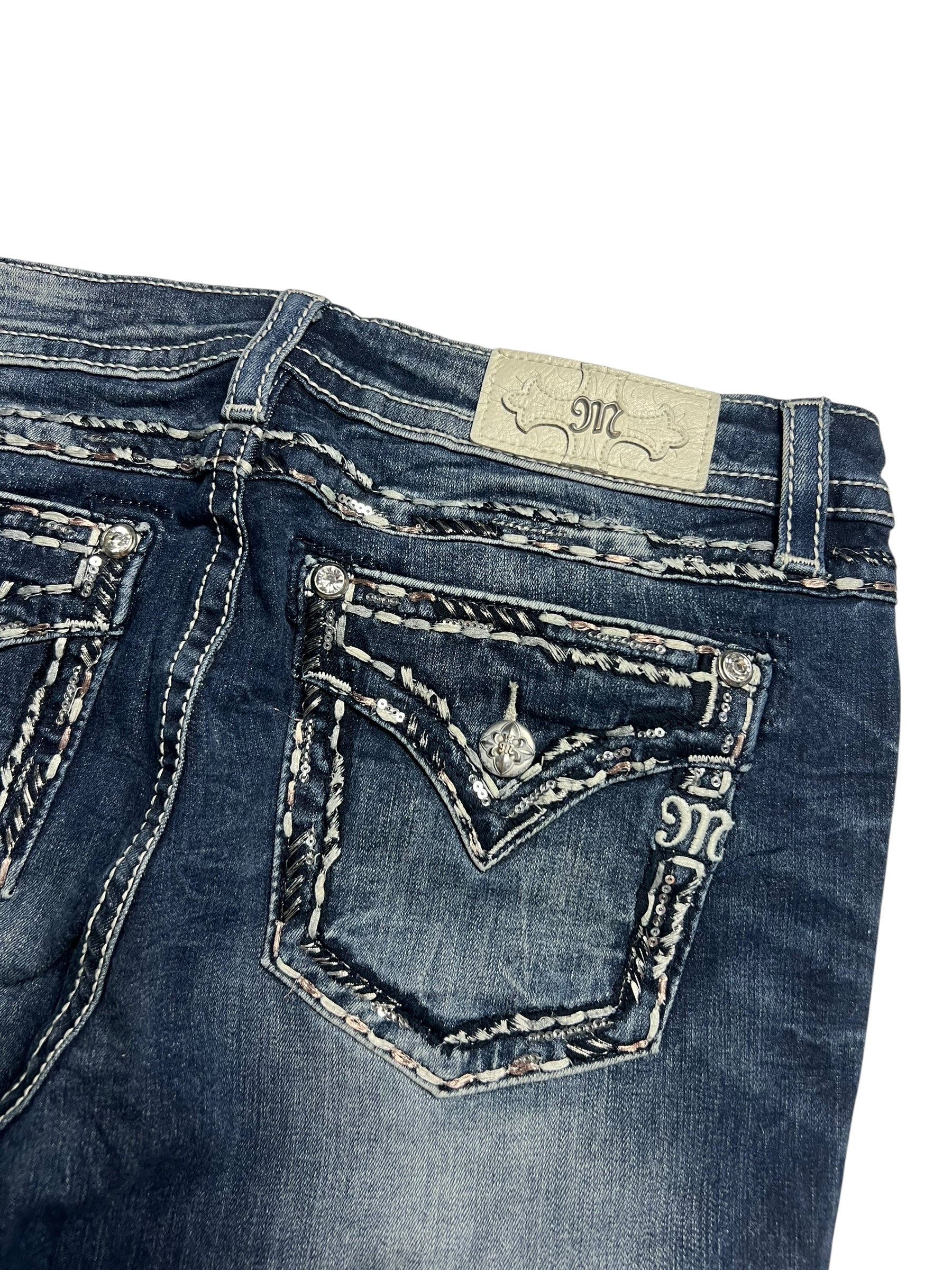 Eine Nahaufnahme der Miss Me Low Rise Boot | 32 (L) Jeans von ZAZA resell. Sie zeigt dunkelblauen Denim, weiße und graue Nähte, Gesäßtaschen mit Klappe und Knöpfen, einen hellen Markenaufnäher aus Leder in der Nähe des Bunds und eine durchsichtige Bundweite zur Größenbestimmung.