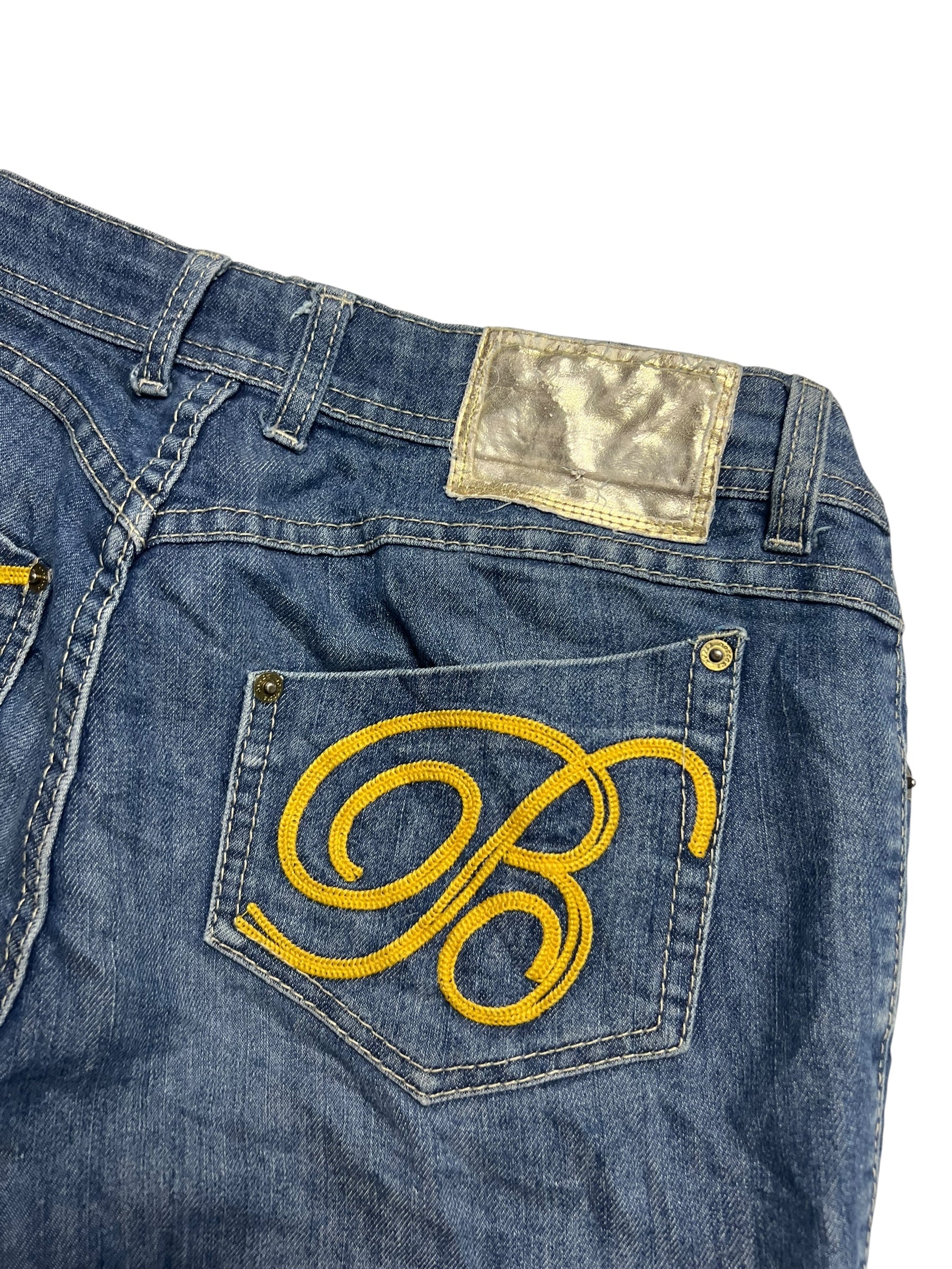 Die Apple Bottoms Mid Rise Boot | M von ZAZA resell ist eine blaue Jeans mit einem gelben gestickten B auf der Gesäßtasche, einem silbernen Metallic-Aufnäher darüber und taillierter Bundweite für einen bequemen Sitz.