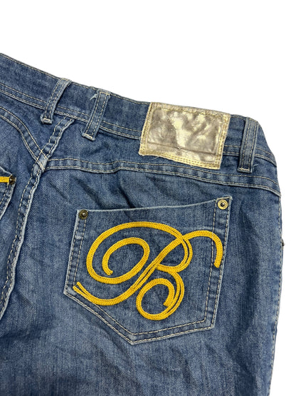 Die Apple Bottoms Mid Rise Boot | M von ZAZA resell ist eine blaue Jeans mit einem gelben gestickten B auf der Gesäßtasche, einem silbernen Metallic-Aufnäher darüber und taillierter Bundweite für einen bequemen Sitz.