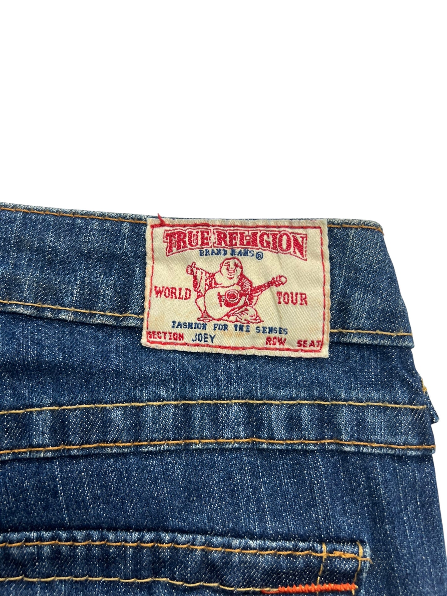 Eine Nahaufnahme der blauen Jeans True Religion Joey - Low Rise Boot von ZAZA resell, Größe 24 (S), mit einem Etikett mit einem Gitarre spielenden Buddha und Angaben wie World Tour, Section Joey, Row Seat.