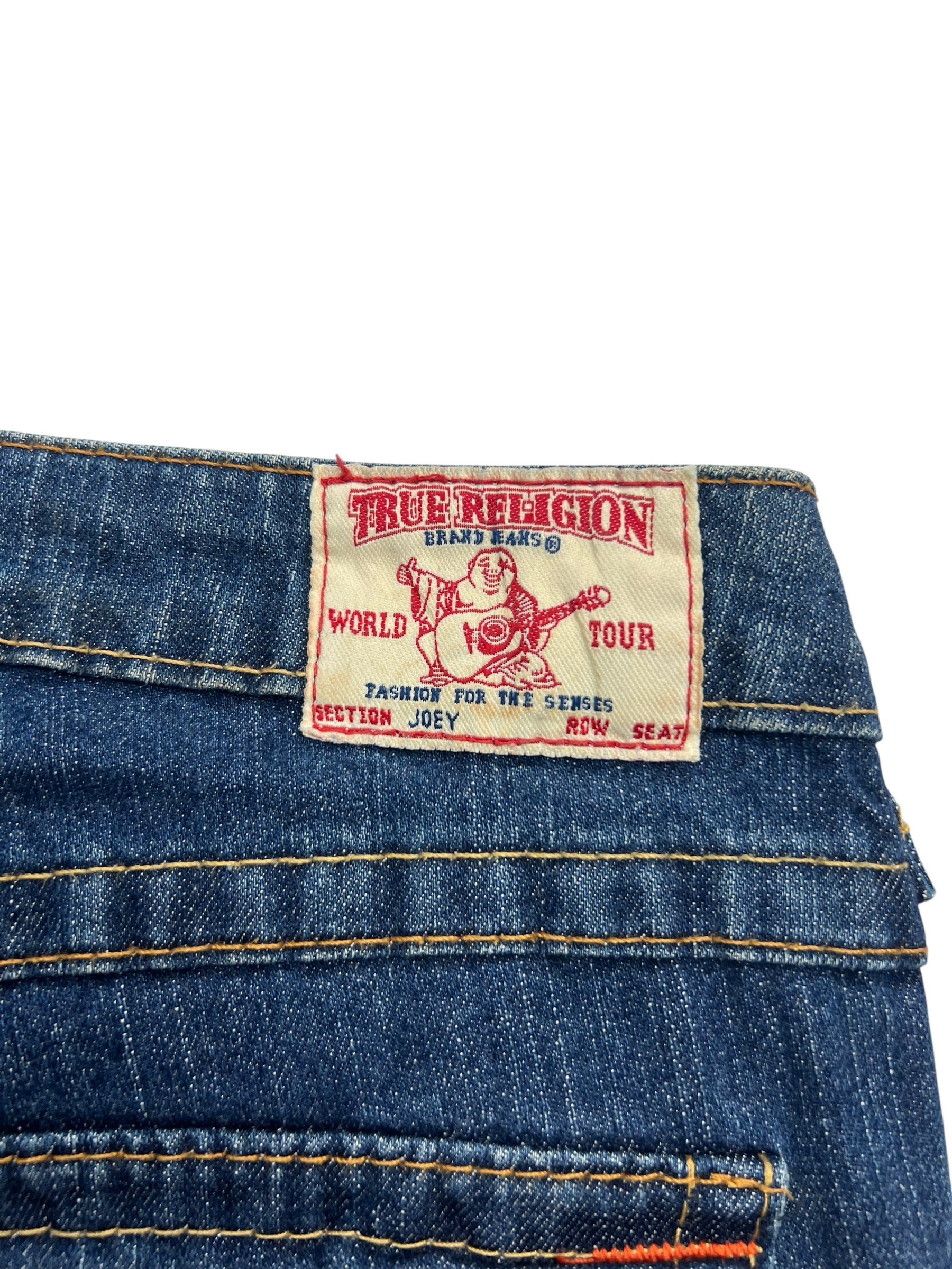 Eine Nahaufnahme der blauen Jeans True Religion Joey - Low Rise Boot von ZAZA resell, Größe 24 (S), mit einem Etikett mit einem Gitarre spielenden Buddha und Angaben wie World Tour, Section Joey, Row Seat.
