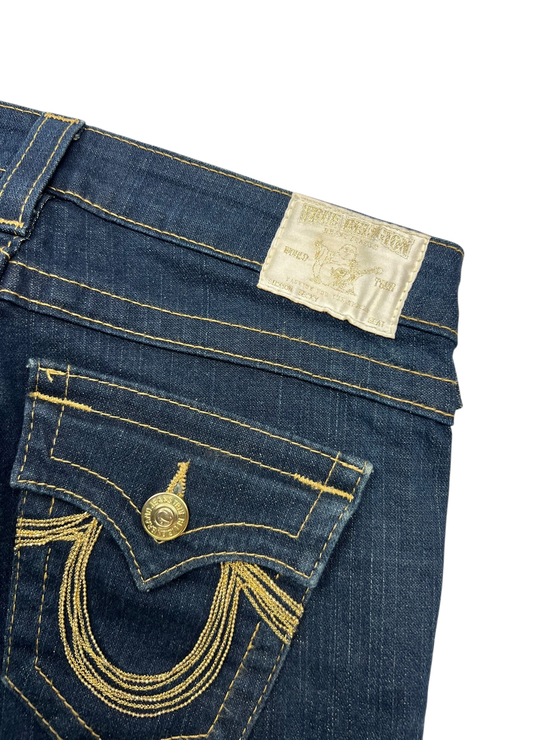 Nahaufnahme der True Religion Becky Low Rise Boot Jeans von ZAZA resell in Größe 26 (S), blauer Denim mit goldenen Nähten, Vordertasche mit Klappe und Knopf und einem weißen Bundetikett. Breite der Taille: 38,5 cm.