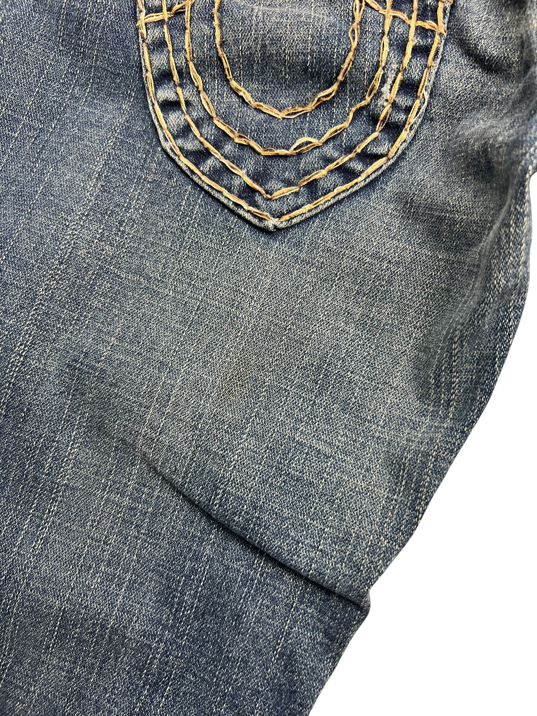 Nahaufnahme der blauen Denim von ZAZA resell's True Religion Joey Super T - Low Rise Boot | 26 (S), die eine Tasche mit gelben Ziernähten und abgenutzter Textur zeigt, die das für die Marke typische Detail hervorhebt.