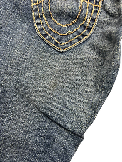 Nahaufnahme der blauen Denim von ZAZA resell's True Religion Joey Super T - Low Rise Boot | 26 (S), die eine Tasche mit gelben Ziernähten und abgenutzter Textur zeigt, die das für die Marke typische Detail hervorhebt.