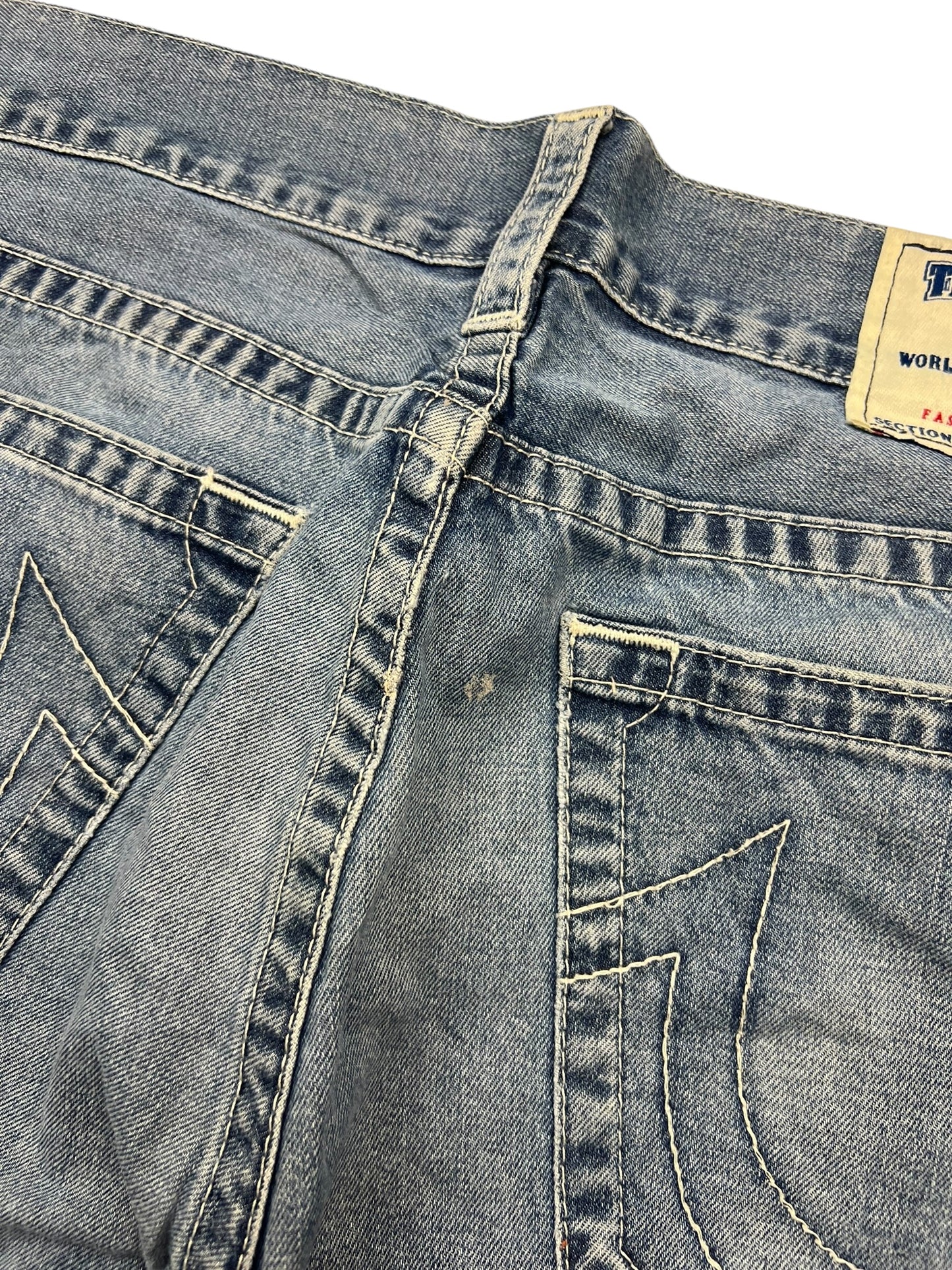 Nahaufnahme der Rückseite einer verblassten blauen True Religion Straight-Jeans (34L) von ZAZA resell, mit abgenutztem Stoff, kleinem Loch in der Nähe der Mittelnaht, dekorativen Taschennähten, teilweisem Bundetikett und subtilen Größendetails.