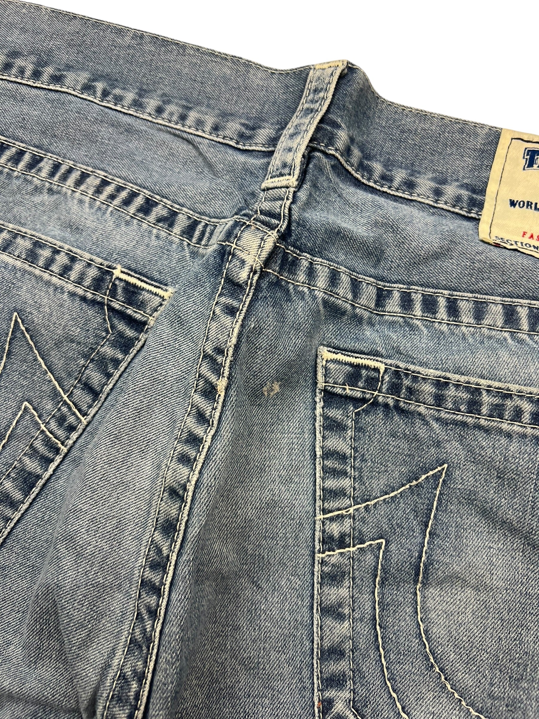 Nahaufnahme der Rückseite einer verblassten blauen True Religion Straight-Jeans (34L) von ZAZA resell, mit abgenutztem Stoff, kleinem Loch in der Nähe der Mittelnaht, dekorativen Taschennähten, teilweisem Bundetikett und subtilen Größendetails.