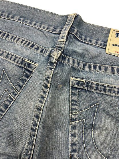 Nahaufnahme der Rückseite einer verblassten blauen True Religion Straight-Jeans (34L) von ZAZA resell, mit abgenutztem Stoff, kleinem Loch in der Nähe der Mittelnaht, dekorativen Taschennähten, teilweisem Bundetikett und subtilen Größendetails.