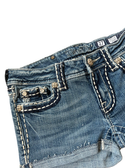 Nahaufnahme der blauen Denim Miss Me Shorts | 27 (S) von ZAZA resell, mit weißen Nähten, Ziernieten, einem sichtbaren Markenetikett am Bund und dem Größenetikett Labelgröße 27 mit Bundweite 35.