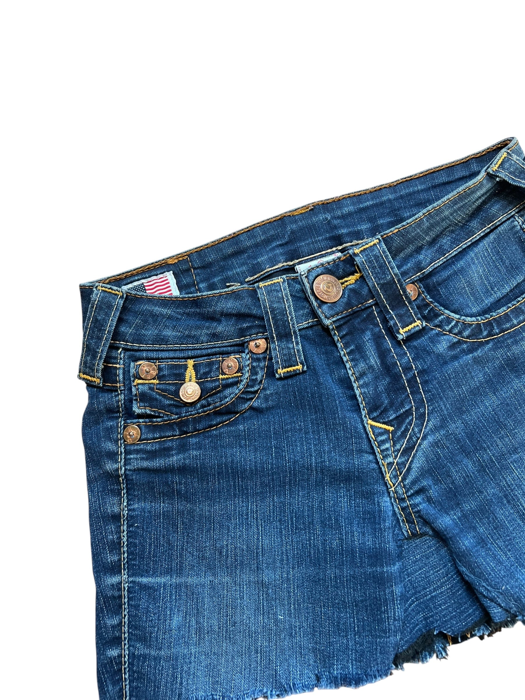 Eine Nahaufnahme des True Religion Skirt | 24 (XS) von ZAZA resell aus blauem Denim mit ausgefransten Säumen, kupferfarbenen Knöpfen, gelben Nähten und Vordertaschen. Außenbeinlänge 27 für eine maßgeschneiderte Rock-inspirierte Passform vor weißem Hintergrund.