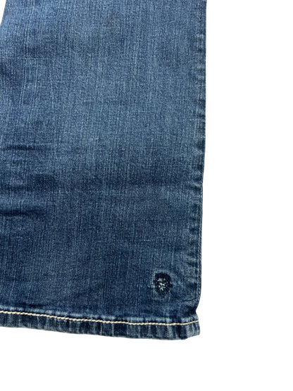 Nahaufnahme der Miss Me Mid Rise Curvy Boot Jeans (28/M) von ZAZA resell. Sie zeigt blauen Denim, einen leicht ausgefransten Saum, einen kleinen kreisförmigen, abgenutzten Fleck in der Nähe der Kante und eine taillierte Bundweite für zusätzlichen Komfort.