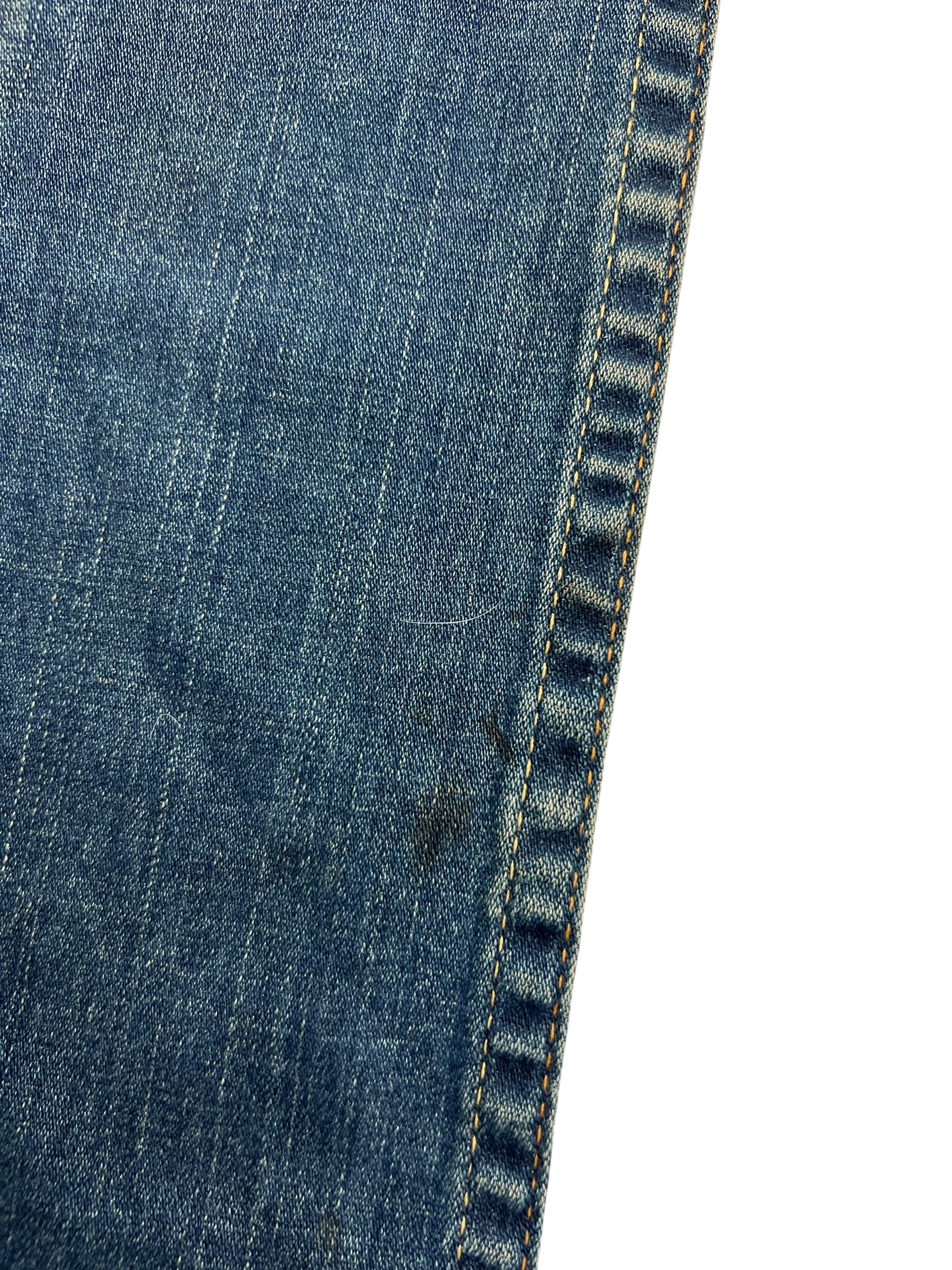 Nahaufnahme von blauem Denim-Stoff mit gelben Nähten und einem kleinen Fleck in der Nähe der Naht. Die True Religion Billy - Low Rise Straight | 24 (S) von ZAZA resell vereint ideale Passform und Qualität für Detailverliebte.