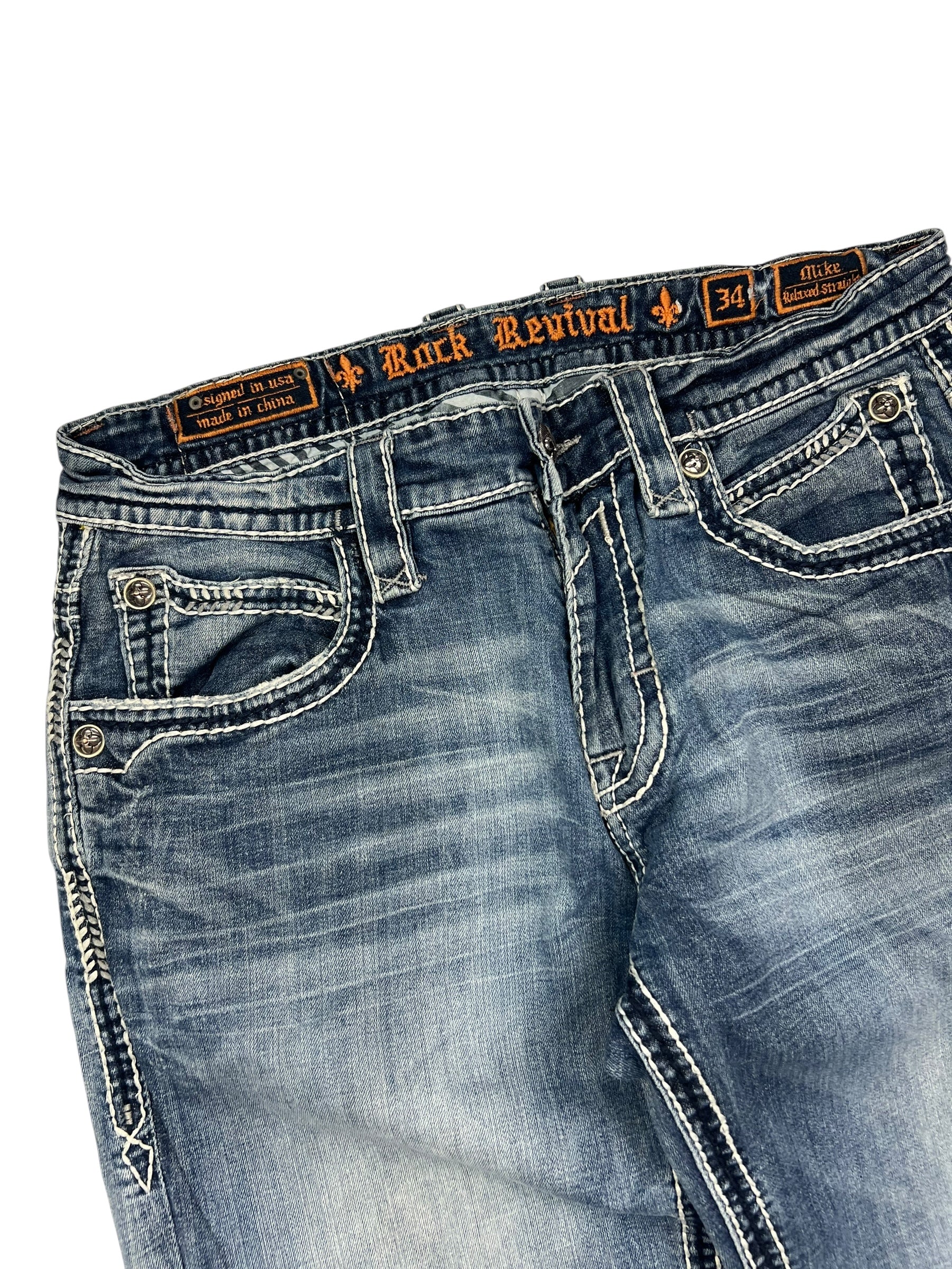 Eine Nahaufnahme der blauen ZAZA resell Rock Revival Relaxed Straight/Baggy Jeans in Größe 34 (M) mit weißen Kontrastnähten, dekorativen Vordertaschen und einem Bundweite-Label am Bund.