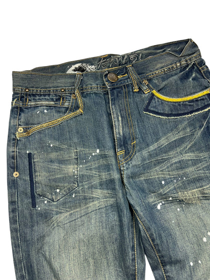 Die Big Train Boot Jeans von ZAZA resell in M/L hat blauen Distressed-Denim, Farbspritzer, einen gelben Reißverschluss an einer Tasche, sichtbares Fading und Whiskering und eine Außenbeinlänge für eine bequeme Passform.