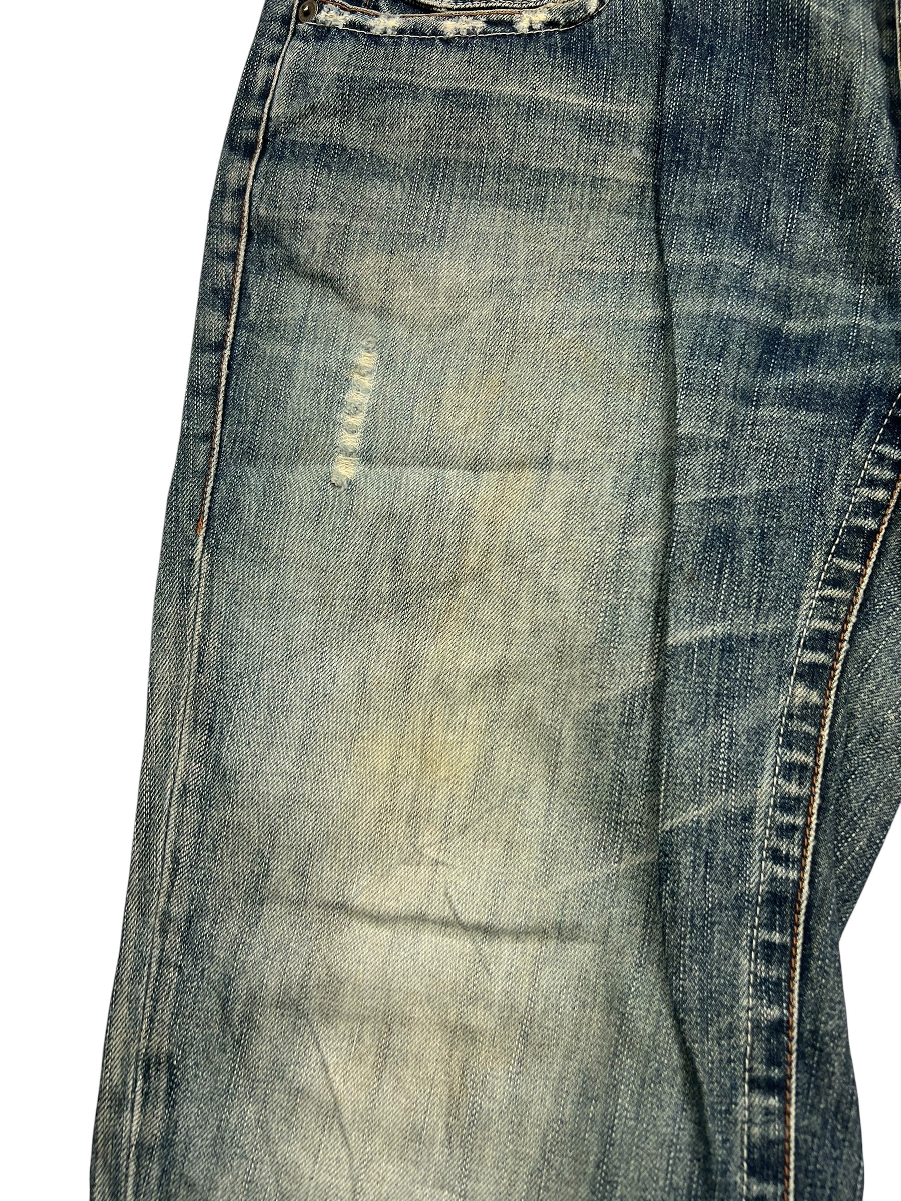 Nahaufnahme der Big Train Boot | L Jeans von ZAZA resell, mit verblasstem blauen Stoff, sichtbaren Nähten, einem reparierten Aufnäher in der Nähe der Tasche und einem klaren Blick auf die Bundweite zur Bewertung des Zustands.