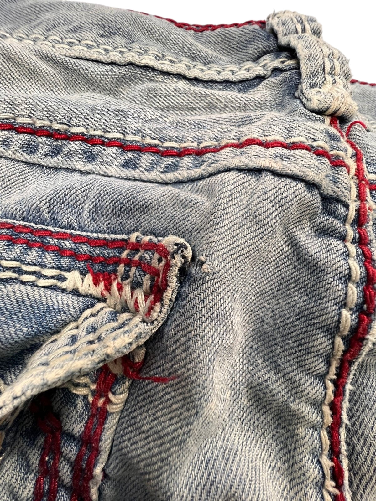 Nahaufnahme der True Religion Mega T Straight-Jeans von ZAZA resell in Größe 34 (M) mit verblasstem blauem Denim, auffälligen roten und weißen Nähten, einem Taschenrand, strukturierter Abnutzung und einem sichtbaren Etikett in der Nähe des Bunds.
