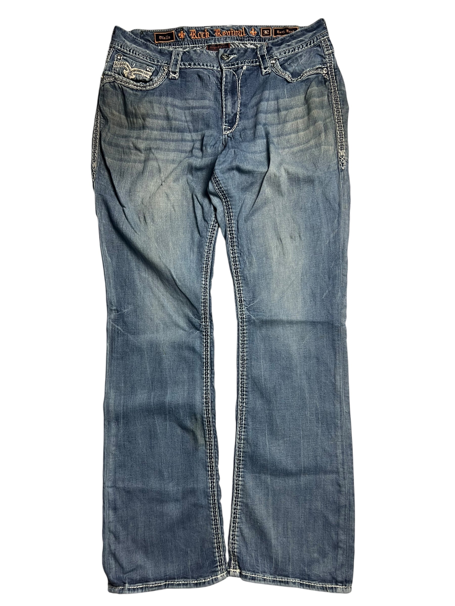Die Rock Revival Mid Rise Boot-Jeans von ZAZA resell (Größe 32 XL, Bundweite 46) hat einen verblichenen blauen Denim, weiße Nähte, Taschen auf der Vorderseite und ein sichtbares Logo-Etikett auf dem Innenbund, das flach auf weißem Grund abgebildet ist.