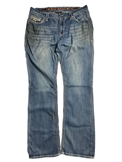 Die Rock Revival Mid Rise Boot-Jeans von ZAZA resell (Größe 32 XL, Bundweite 46) hat einen verblichenen blauen Denim, weiße Nähte, Taschen auf der Vorderseite und ein sichtbares Logo-Etikett auf dem Innenbund, das flach auf weißem Grund abgebildet ist.