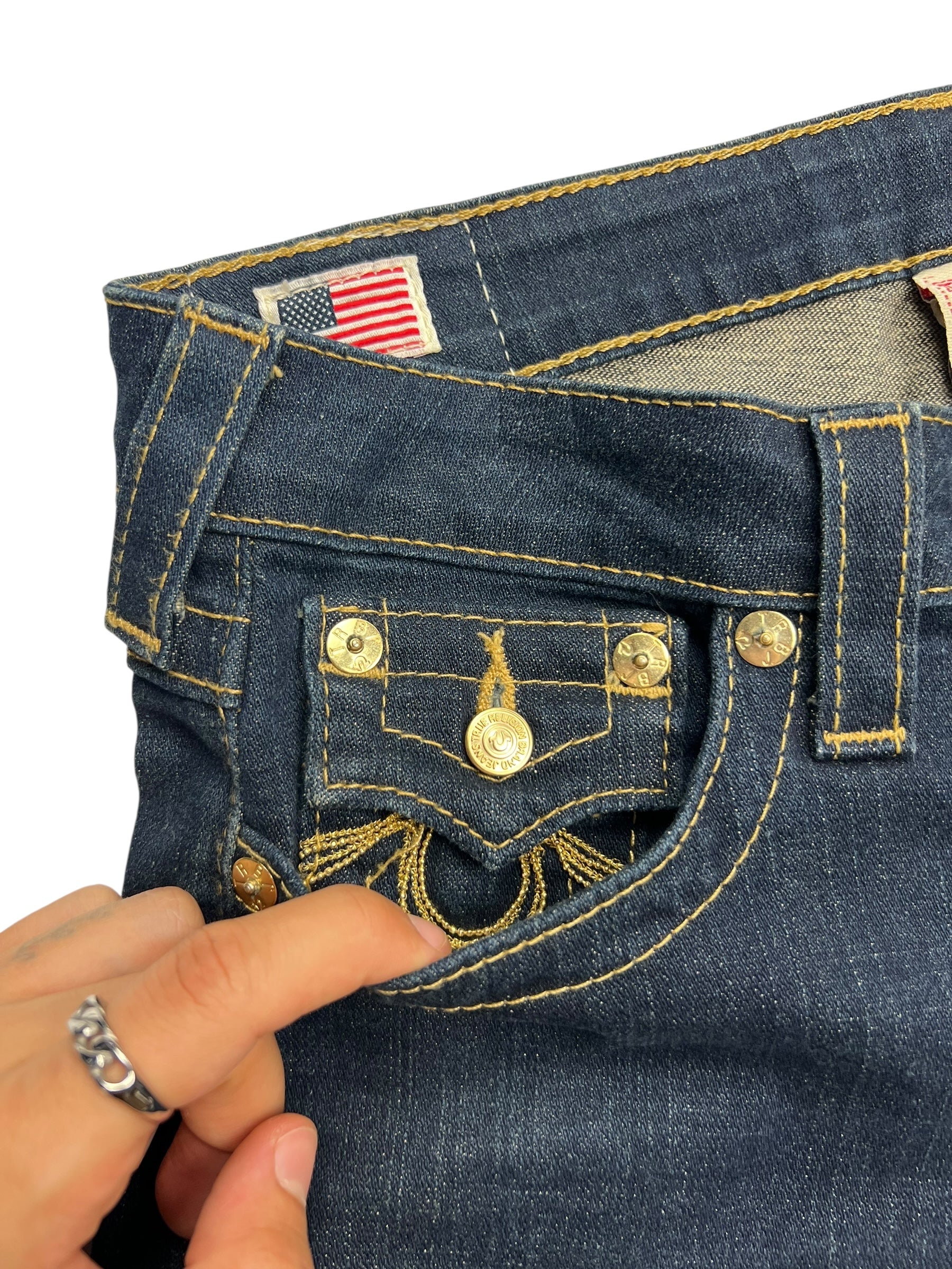 Eine Hand mit einem silbernen Ring öffnet die kleine Vordertasche der True Religion Becky Low Rise Boot Jeans von ZAZA resell, Größe 26 (S), und enthüllt goldene Nähte, Metallnieten und ein Schild mit der amerikanischen Flagge am Bund.