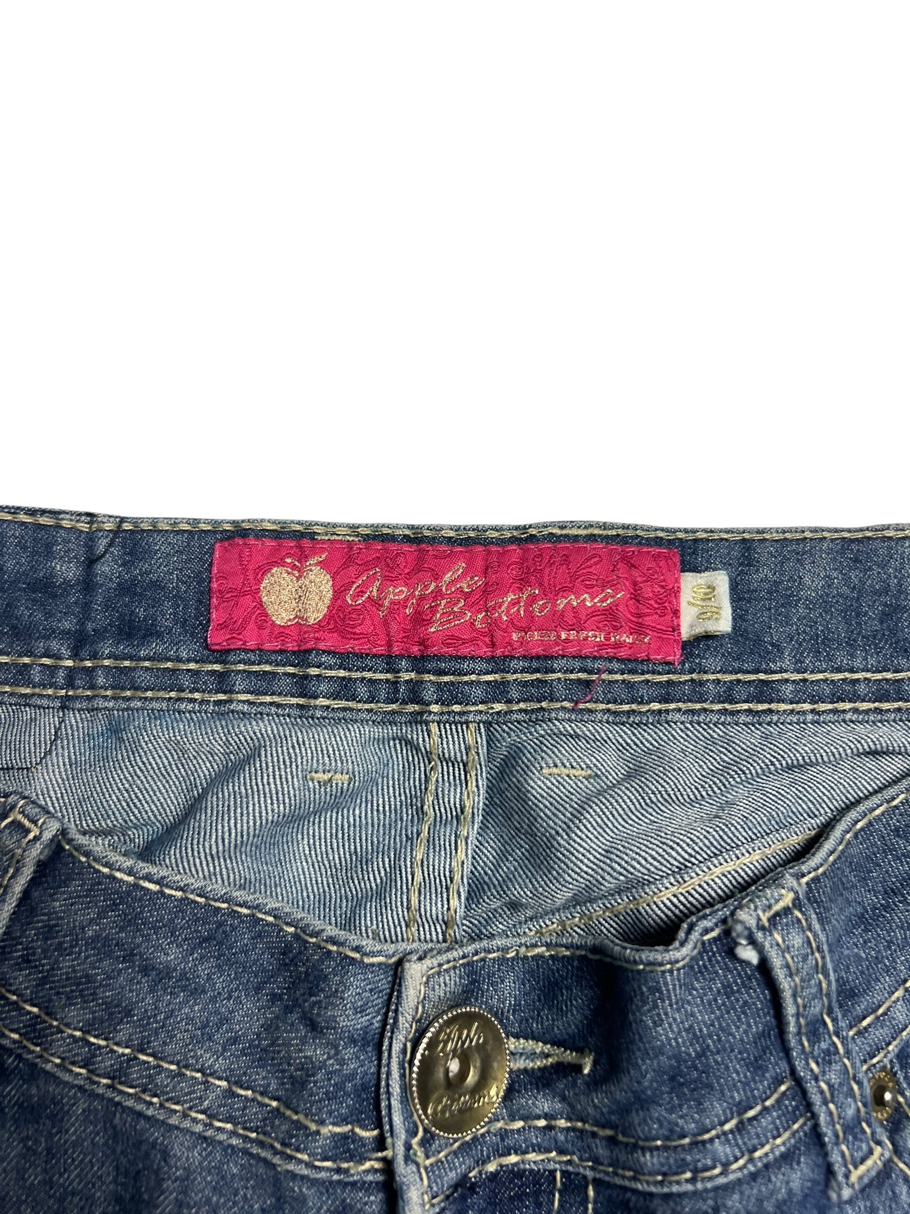 Nahaufnahme der Apple Bottoms Mid Rise Boot Jeans von ZAZA resell. Mit blauem Denim, rosa Apfel-Symbol-Etikett über dem Bund, sichtbarem Knopf, Nahtdetails und klarer Bundweite für einen klassischen Look.