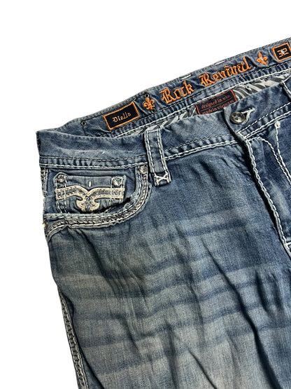 Eine Nahaufnahme der Rock Revival Mid Rise Boot Jeans (32/XL) von ZAZA resell hebt die detaillierten Nähte, die Gürtelschlaufe, die orangefarbene Markenstickerei am Bund und den leicht verblichenen, abgenutzten Look hervor.