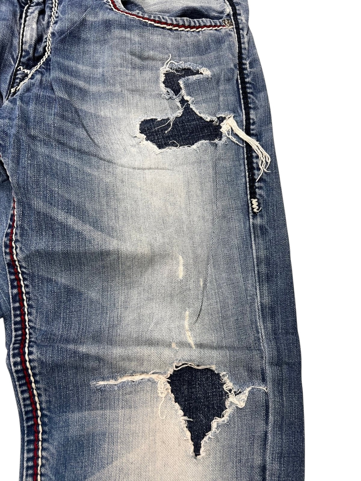 Nahaufnahme der True Religion Mega T Straight-Jeans von ZAZA resell in Größe 34 (M) mit starken Ausfransungen und Rissen an Oberschenkel und Knie, sichtbarem dunklem Unterstoff, detaillierten Nähten und rot-weiß-blauen Seitenstreifen.