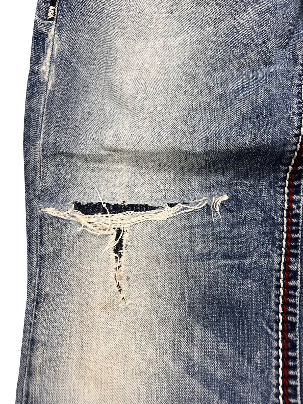 Nahaufnahme der True Religion Mega T Straight Jeans von ZAZA resell in Größe 34 (M). Die Jeans ist aus blauem verblichenem Denim mit einem großen horizontalen Riss, ausgefransten Kanten, die weiße Fäden erkennen lassen, und einem aufgenähten rot-weißen Streifen an der rechten Naht.